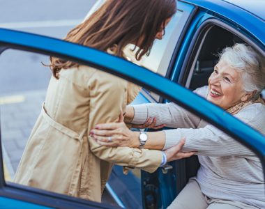 A joyful elderly woman is being helped out of a blue car by a caregiver. The scene conveys warmth, support, and care, highlighting the bond between different generations.